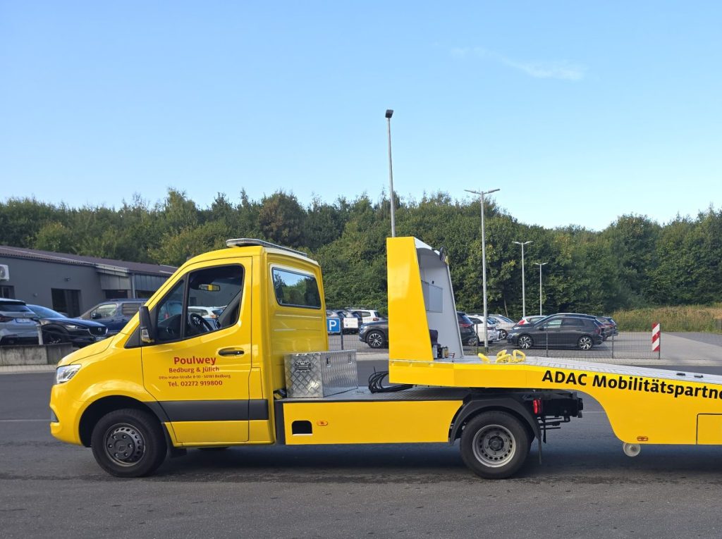 Gelber Abschleppwagen mit einer flachen Anhängerplattform steht auf einer Straße.