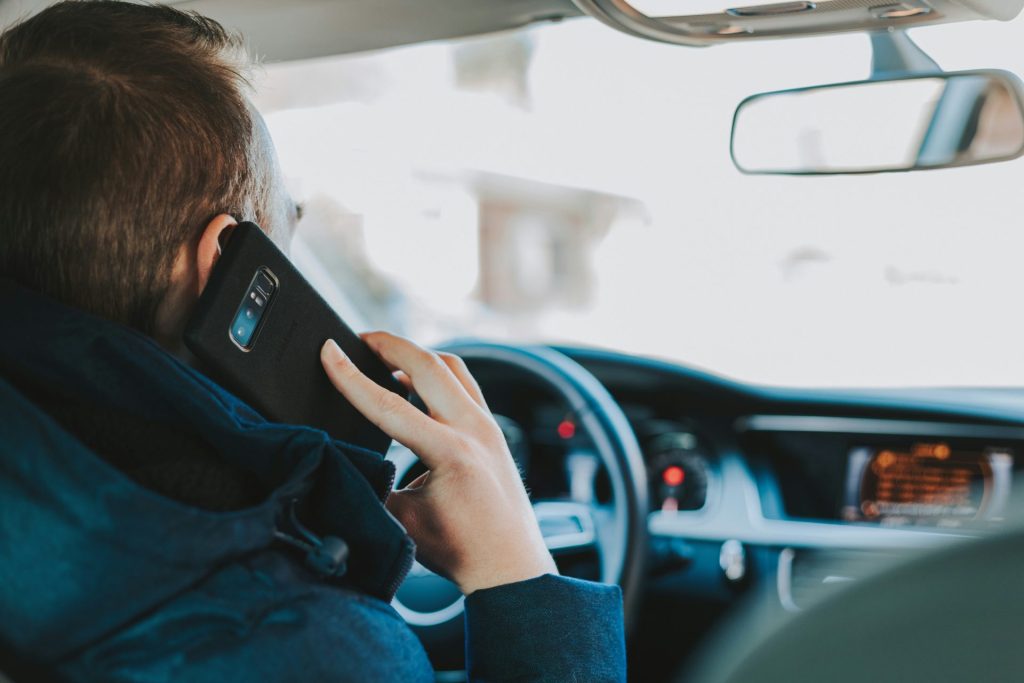 Ein Mann sitzt auf dem Autositz vor dem Lenkrad und hält ein Telefon in der Hand.
